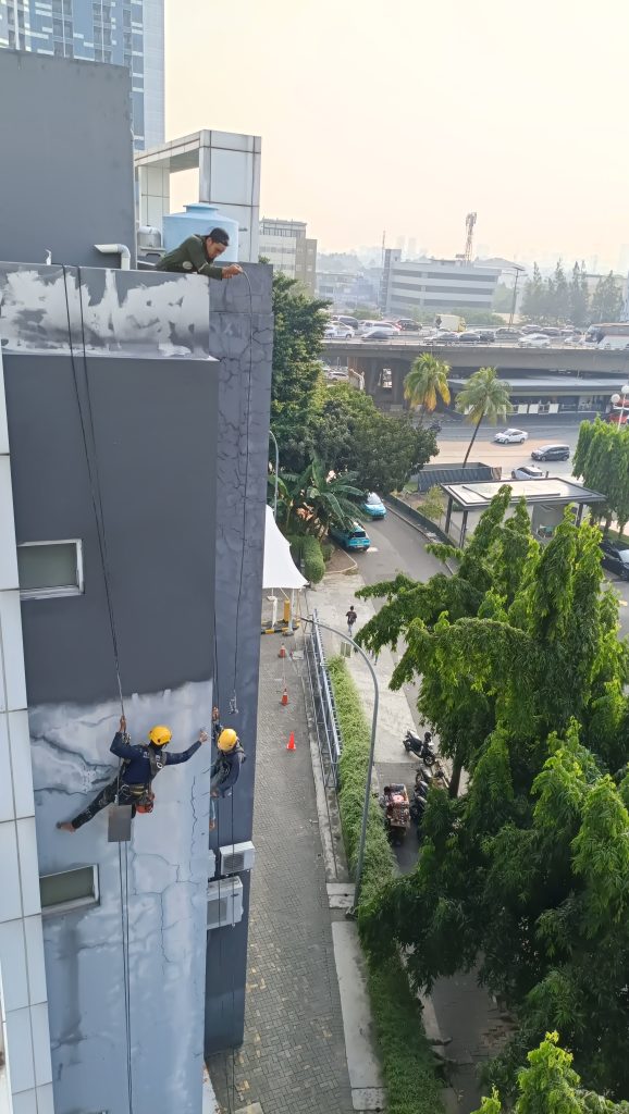 Dua pekerja menggunakan peralatan rope access untuk mengerjakan waterproofing pada dinding luar gedung tinggi